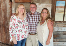 Three people stand smiling in front of a rustic wooden wall. Two women stand beside a man in a warm and friendly atmosphere.