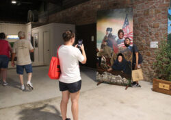Visitors explore a brick-walled museum as a woman photographs a historical mural depicting diverse figures, an American flag, and a river scene.