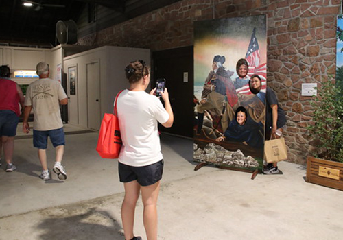 Visitors explore a brick-walled museum as a woman photographs a historical mural depicting diverse figures, an American flag, and a river scene.