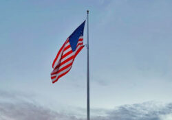 American flag flies on a flag pole in the clear blue sky. Some clouds float in the background.