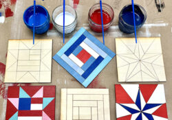 Six wooden tiles with quilt patterns, some painted red, white, and blue, lie on a surface. Nearby, jars with paints and brushes.