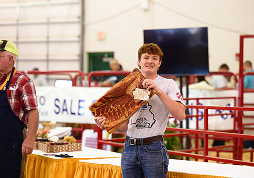 A young person proudly holds a slab of bacon with a prize tag in a bustling indoor venue. He smiles broadly, conveying a sense of achievement.