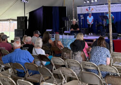 Audience seated under a tent watches a man speaking on a stage with a woman nearby. A backdrop reads "Touchstone Energy Cooperatives of Missouri."