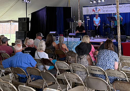 Audience seated under a tent watches a man speaking on a stage with a woman nearby. A backdrop reads "Touchstone Energy Cooperatives of Missouri."