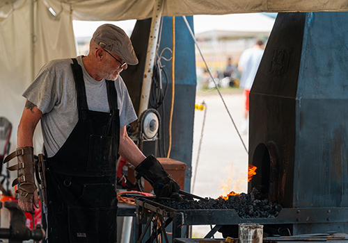 A blacksmith in overalls and a cap works at a forge, focusing intently on fiery coals under a tent. The scene conveys dedication and craftsmanship.