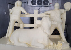 A butter sculpture named "The Great American Past Time" showing two kids facing a fence and one is holding a baseball glove like they are going to catch a ball. A cow sits behind them with a bucket of popcorn laying next to him.