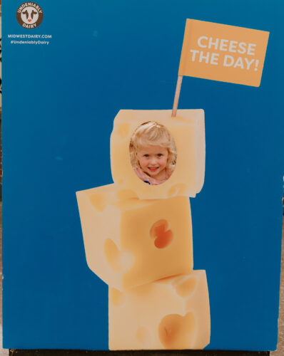 Kid standing behind a photo backdrop that is blue with cheese stacked on it. The top cheese has a hole for her face and a flag that says "cheese the day"