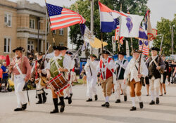 A group of people in historical American Revolutionary War attire marches in a parade, carrying flags, including the American flag, with a festive atmosphere.