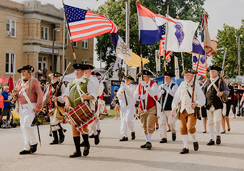A group of people in historical American Revolutionary War attire marches in a parade, carrying flags, including the American flag, with a festive atmosphere.