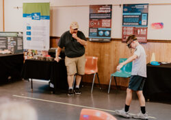 A boy wearing simulation goggles walks along a line in a room, supervised by a man. Educational posters and displays about driving safety are visible.