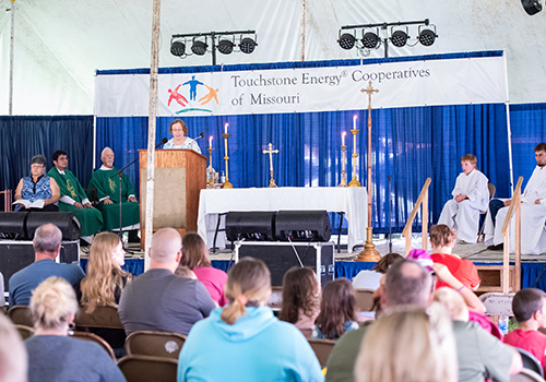 People gather under a tent for a service by Touchstone Energy Cooperatives of Missouri. A woman speaks at a podium; clergy and attendees listen.