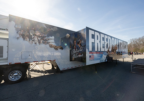 A large truck trailer with "Freedom Truck" written boldly on its side, adorned with a patriotic mural. The scene is sunlit and set in an open area.