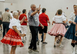 A group of people square dancing indoors, wearing colorful skirts and casual attire. The atmosphere is lively and engaging, with a cheerful vibe.