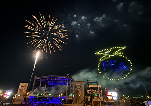 Firework and drone show light up the night sky over an outdoor stage. The drones form the words "FFA" in blue with the yellow FFA emblem surrounding it. Bright, festive atmosphere with glowing lights.
