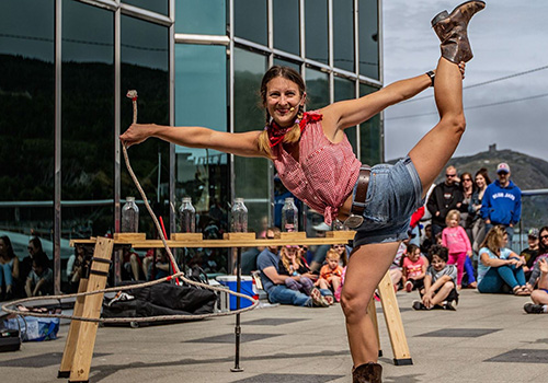 Performer in a Western-themed outfit balances on one leg during an outdoor show. Audience seated in background. Joyful and lively atmosphere.