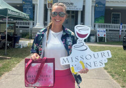 A woman stands in front of a building on a sidewalk holding a Missouri Wine bag and sign