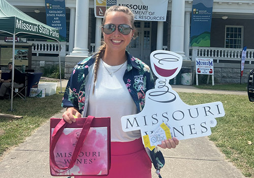 A woman stands in front of a building on a sidewalk holding a Missouri Wine bag and sign