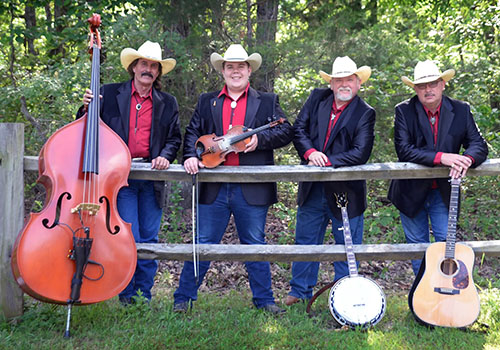 Four men in cowboy hats and black jackets stand by a wooden fence, holding string instruments outdoors. They appear cheerful and relaxed.