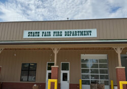 State Fair Fire Department building with a beige facade, green sign, garage door, window, and two yellow bollards. Sky with light clouds above.