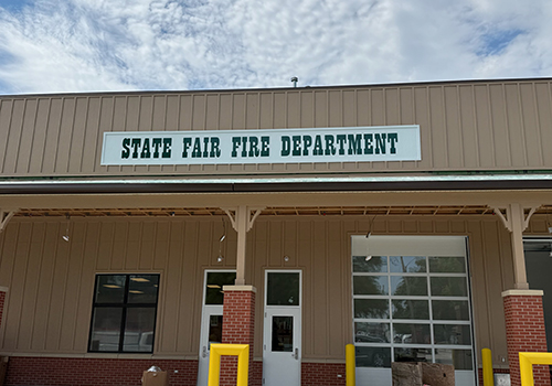 State Fair Fire Department building with a beige facade, green sign, garage door, window, and two yellow bollards. Sky with light clouds above.