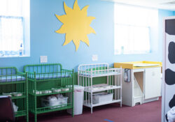 Bright nursery room with blue walls features a large sun decoration. Green and white cribs line the wall, and a yellow changing table is nearby.