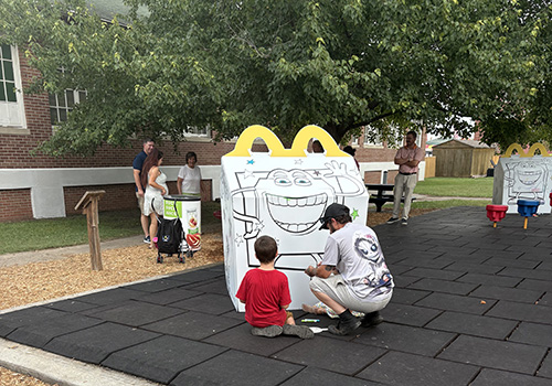 Children and adults engage in outdoor coloring at a playground. A man and a boy focus on an oversized Happy Meal Box. The scene is lively and communal.