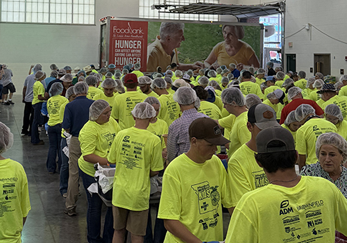 Kids in bright yellow shirts stand in a building with a Feeding Missouri trucki in the backgrounds. The enviorment suggests food packing.