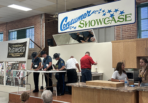 Police officers in uniform participate in a cooking demonstration at a Consumer Showcase event. Observers watch as they work on tall countertops.