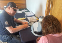 A man wearing a cap and sunglasses assists a woman using a vision screening device at a table. Papers and tissues are on the table, creating a focused atmosphere.