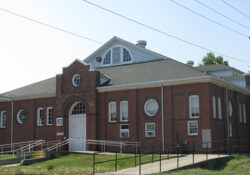A historic brick building with arched windows and a gabled roof. It features circular windows, a ramp, and a path, conveying a vintage charm.