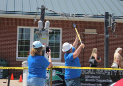 Two people in blue shirts demonstrate electrical safety by handling power line equipment. Yellow caution tape marks the area. A brick building is in the background.