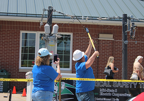 Two people in blue shirts demonstrate electrical safety by handling power line equipment. Yellow caution tape marks the area. A brick building is in the background.