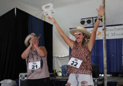 Two women wearing hats joyfully celebrate on stage at a contest. One claps while the other raises her arms, holding a fan. Both wear numbered tags.