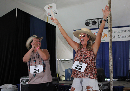 Two women wearing hats joyfully celebrate on stage at a contest. One claps while the other raises her arms, holding a fan. Both wear numbered tags.