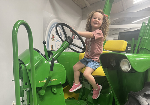 A smiling child with curly hair sits happily on a bright green tractor in an indoor setting, creating a playful and joyful atmosphere.