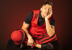 A person sits thoughtfully against a dark background, wearing a red and black striped outfit with matching hat. The mood is playful and whimsical.
