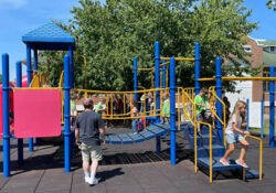 Children play at a playground with adult supervison. The playground has blue and yellow climbing structures and slides. A sunny day with a large tree in the background.