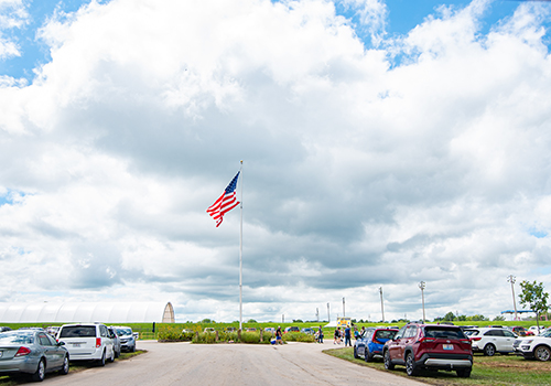 A flag stands tall in the middle of a round about with cars parked in the grass along the road and a cloudy sky is in the background.