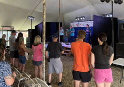 Audience gathered under a tent watches musicians perform on stage. The scene conveys a lively atmosphere with people casually dressed, enjoying the music.