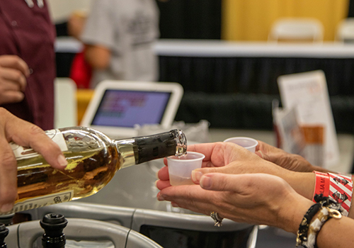 Hands pour wine from a bottle into small plastic cups at a tasting event. The scene is lively, with people in the background and visible event wristbands.