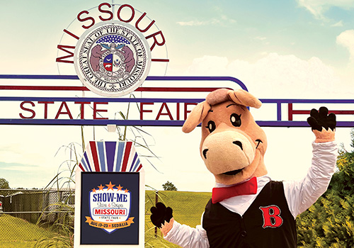 Fair Mascot, Bartholomule, in a tuxedo and red bow tie waves cheerfully in front of a the Missouri State Fair Main Gate Tribute. A fair poster is visible below the sign.