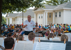 Man stands as conductor in front of a band outdoors with people standing in the background