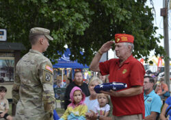 A veteran in a red cap salutes a soldier in camouflage uniform while holding a folded flag. Onlookers, including children, observe respectfully.