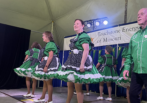 A group of dancers in green outfits with lace, performing on stage under a tent. The sign behind them reads "Touchstone Energy of Missouri."