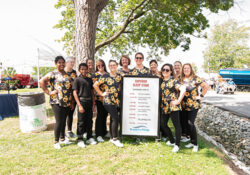 A group of people poses cheerfully around a sign listing event times on the "Superior Sleep Stage." They wear matching sunflower-themed outfits outdoors.