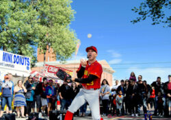 Street performer in a red and yellow costume juggles under a clear blue sky. A crowd watches, with signs for mini donuts and ice cream visible. Vibrant and lively atmosphere.