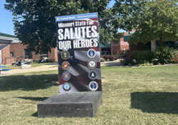 A photo station at the Missouri State Fair reads "Salutes Our Heroes" with military emblems. It stands on grass under a tree, with a brick building nearby.