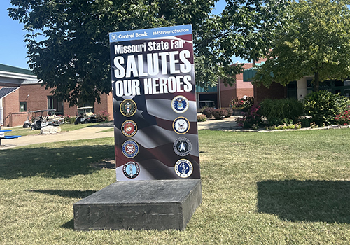 A photo station at the Missouri State Fair reads "Salutes Our Heroes" with military emblems. It stands on grass under a tree, with a brick building nearby.