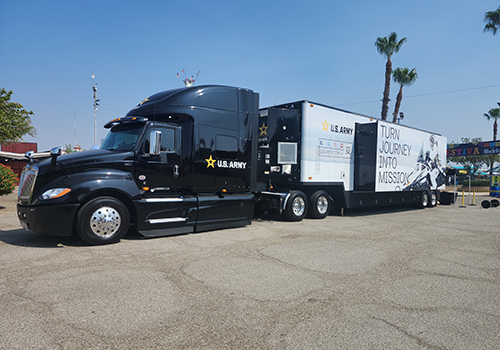 Black U.S. Army semi-truck with attached trailer parked on sunny day. Trailer has "Turn Journey into Mission" slogan and palm trees in background.