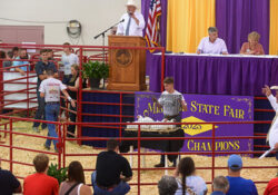 Missouri State Fair livestock auction scene with an auctioneer speaking, audience seated, and a person presenting an animal in a cage. Purple and yellow championship banners in the background.